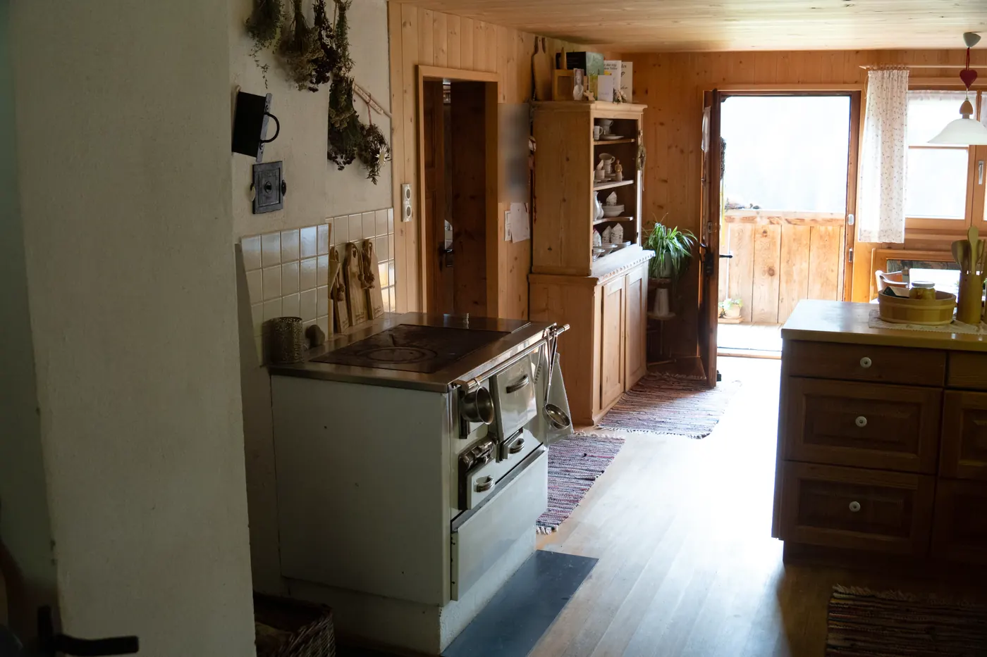 Küche mit weißem Holzherd, getrockneten Kräuterbüscheln an der Wand und offener Tür ins Freie