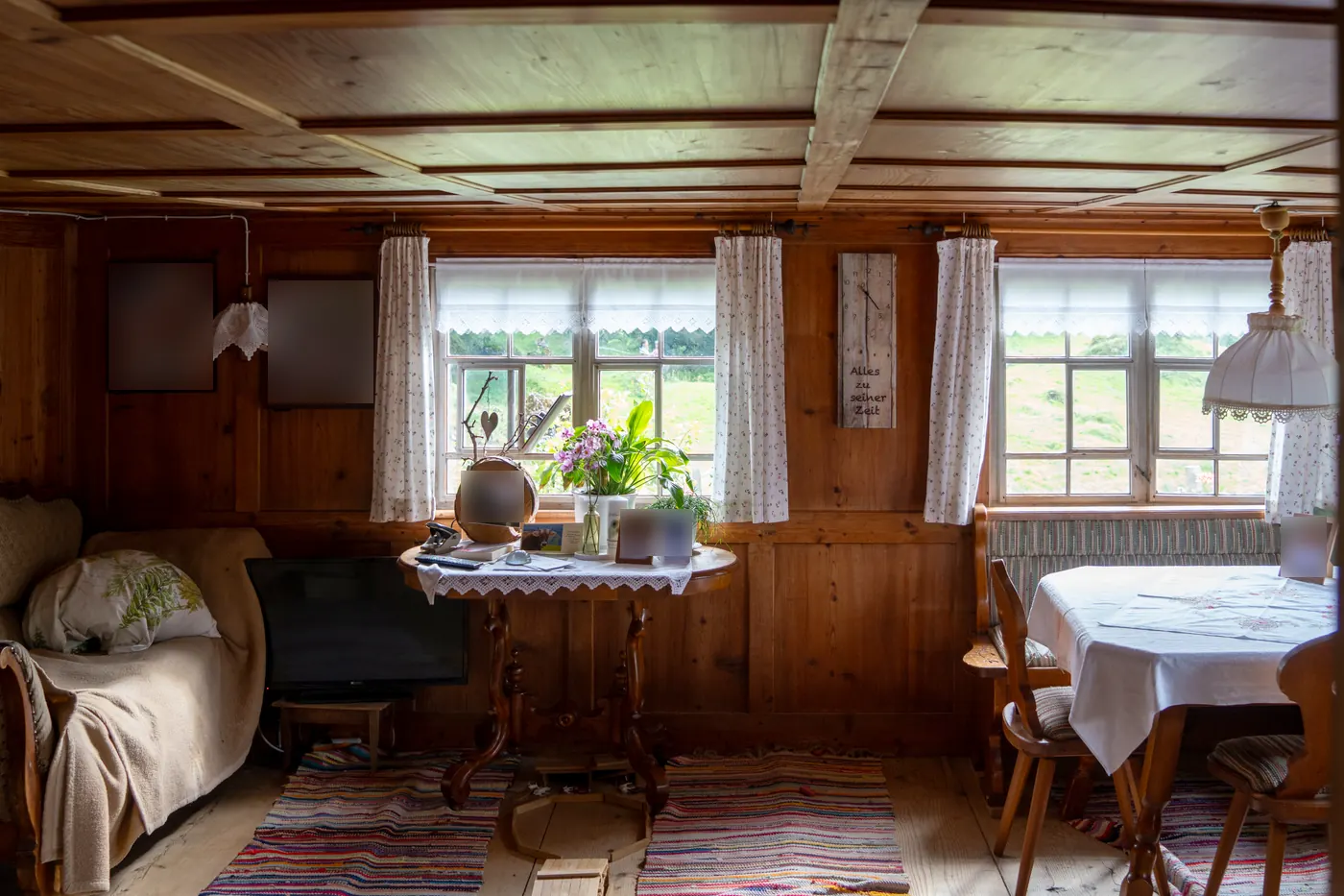 Weiterer Blick in die Stube mit Sofa, Fernseher, Fotocollagen an der Wand und einer Holzuhr mit der Aufschrift Alles zu seiner Zeit