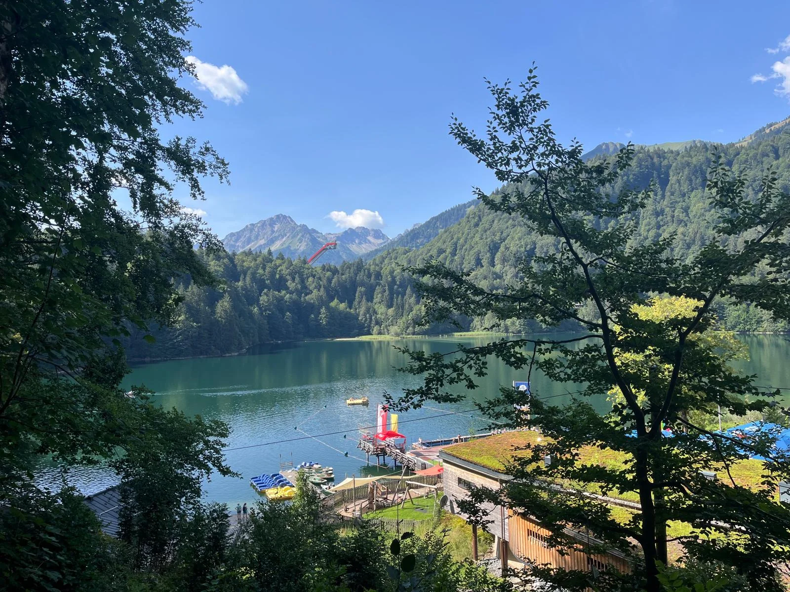Blick durch Bäume auf den Freibergsee mit Naturbad, Steg, Tretbooten und Spielturm im Wasser, dahinter die rote Heini-Klopfer-Skiflugschanze und die Allgäuer Berge