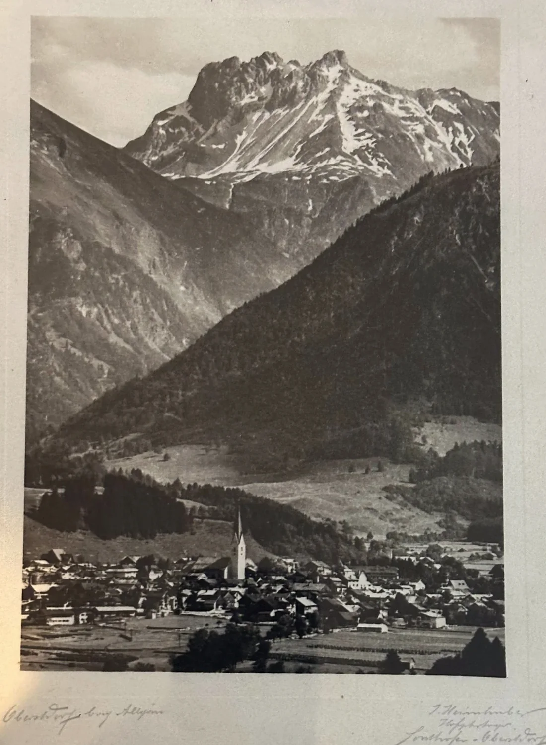 Alte Schwarzweiß-Fotografie: das Dorf Oberstdorf im Talboden mit spitzem Kirchturm, dahinter eine bewaldete Flanke und der zerklüftete Kratzer mit Schneeresten in den Rinnen