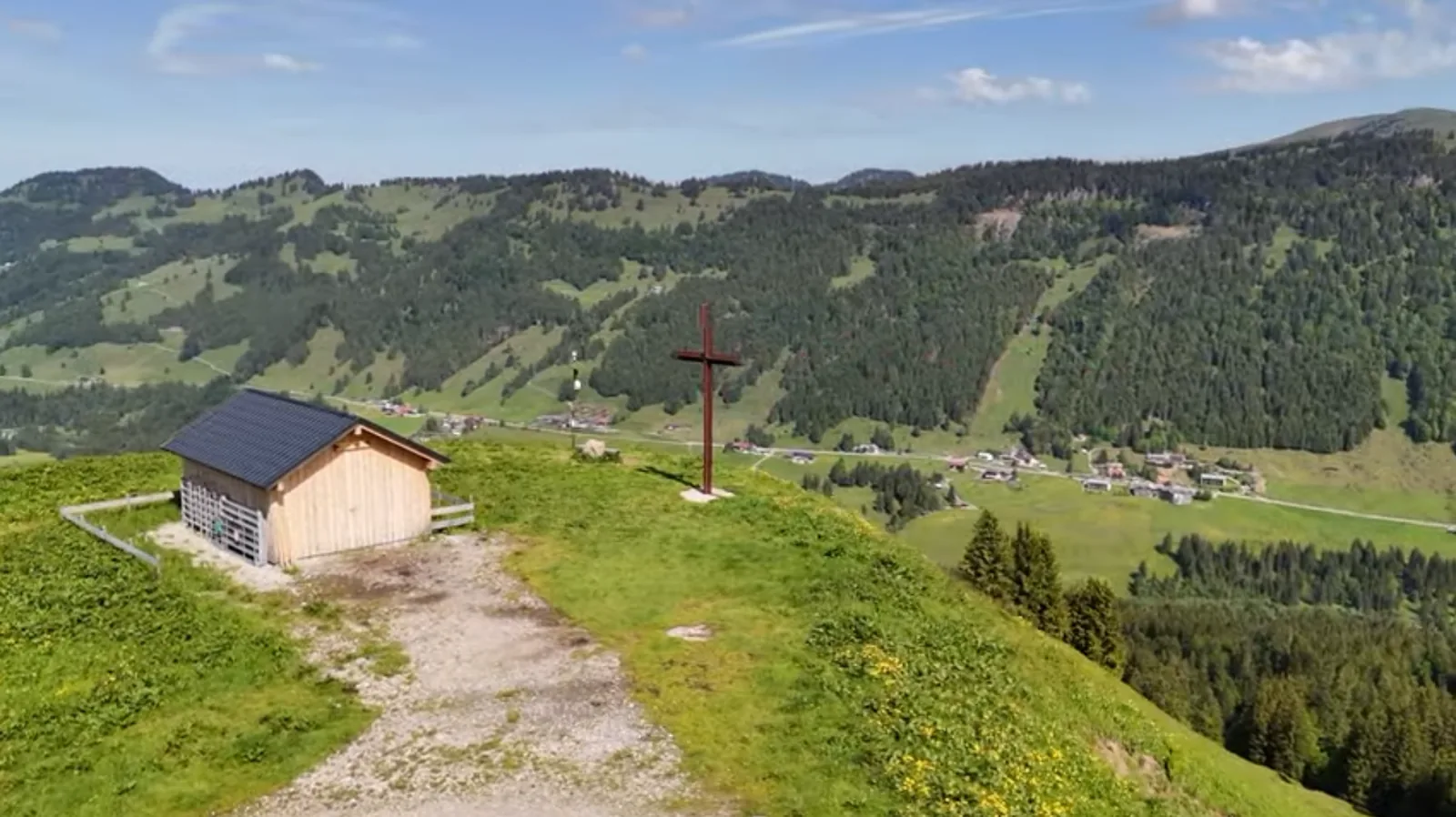 Das Gipfelkreuz auf dem Gelbhansekopf bei Tag: rechts das dunkle Holzkreuz auf einem Betonsockel, links eine Holzhütte, dahinter das Balderschwanger Tal und die bewaldeten Rücken der Nachbarberge