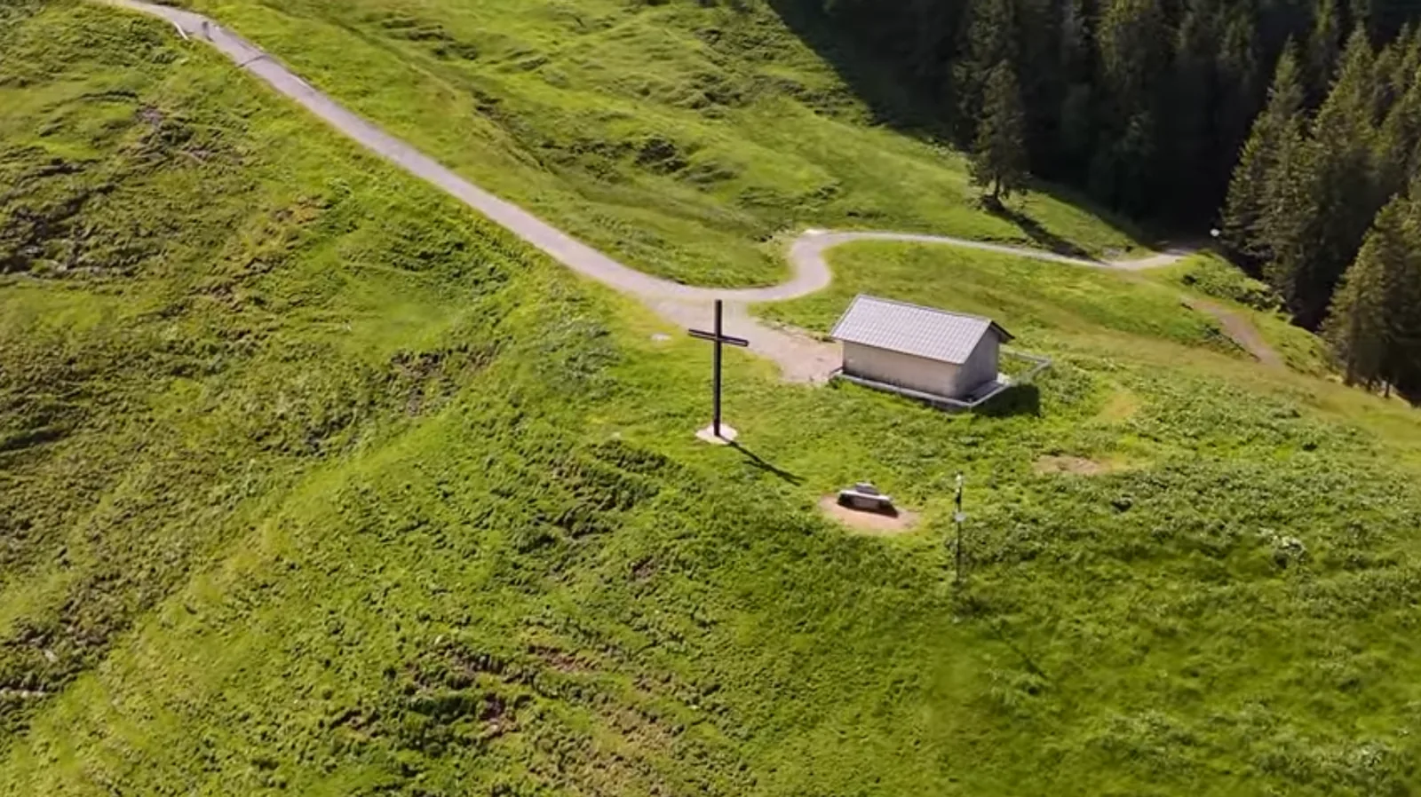 Blick von oben auf den Rücken des Gelbhansekopfs: ein geschotterter Weg führt an Kreuz und Hütte vorbei, ringsum Bergwiese und Zwergsträucher, rechts der Wald