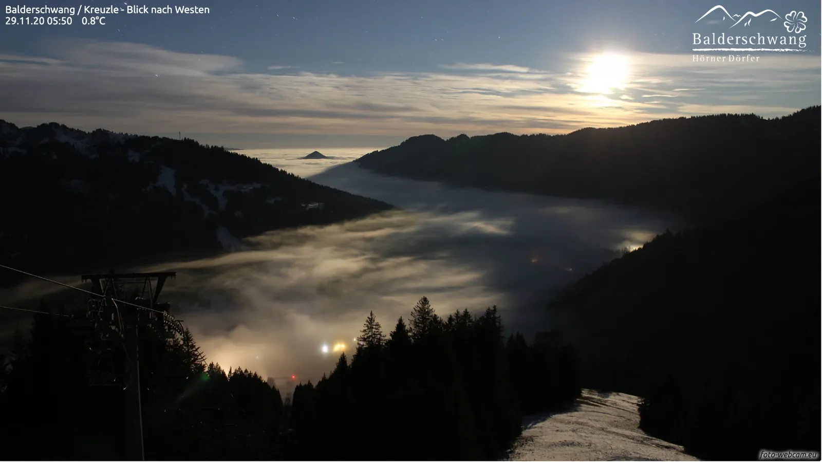 Vollmond über einem Nebelmeer, das das Tal bis zu den Kämmen füllt, im Nebel schimmern die Lichter des Dorfes