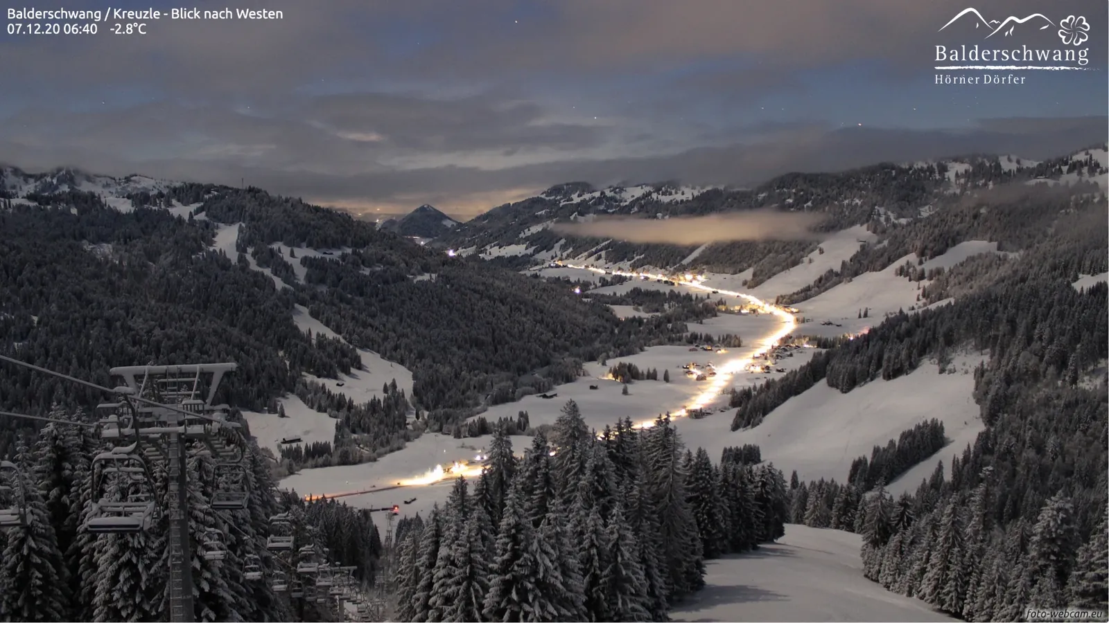 Frühmorgens im Winter: frisch verschneite Bäume, die beleuchtete Straße zieht sich durchs Tal, eine Nebelbank über dem Dorf