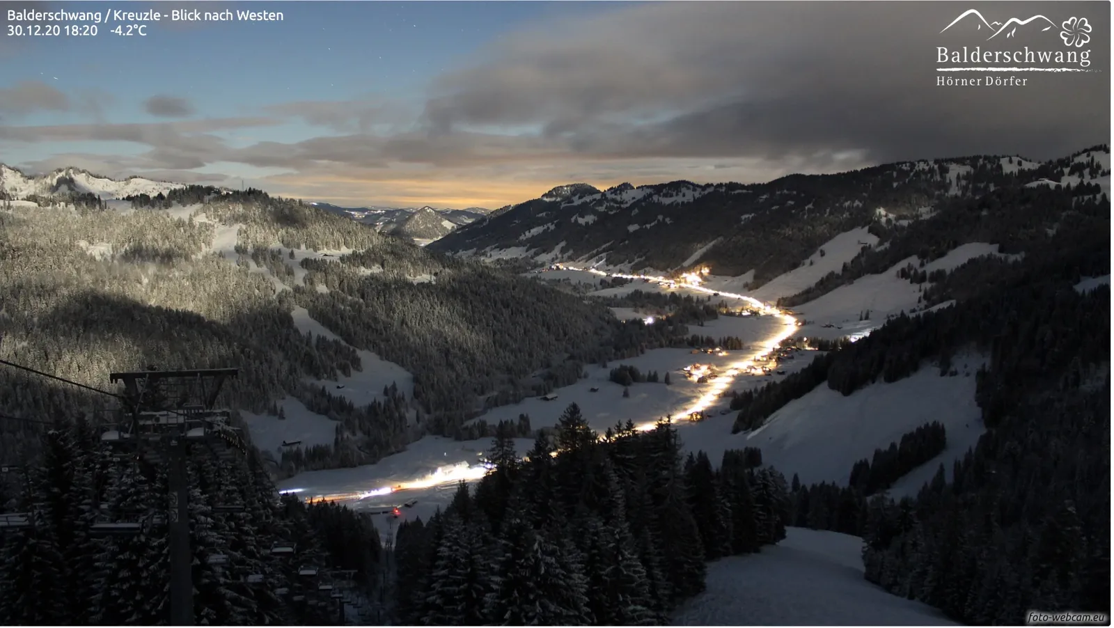 Dämmerung über dem verschneiten Tal, die beleuchtete Straße schlängelt sich als helles Band durch das Dorf
