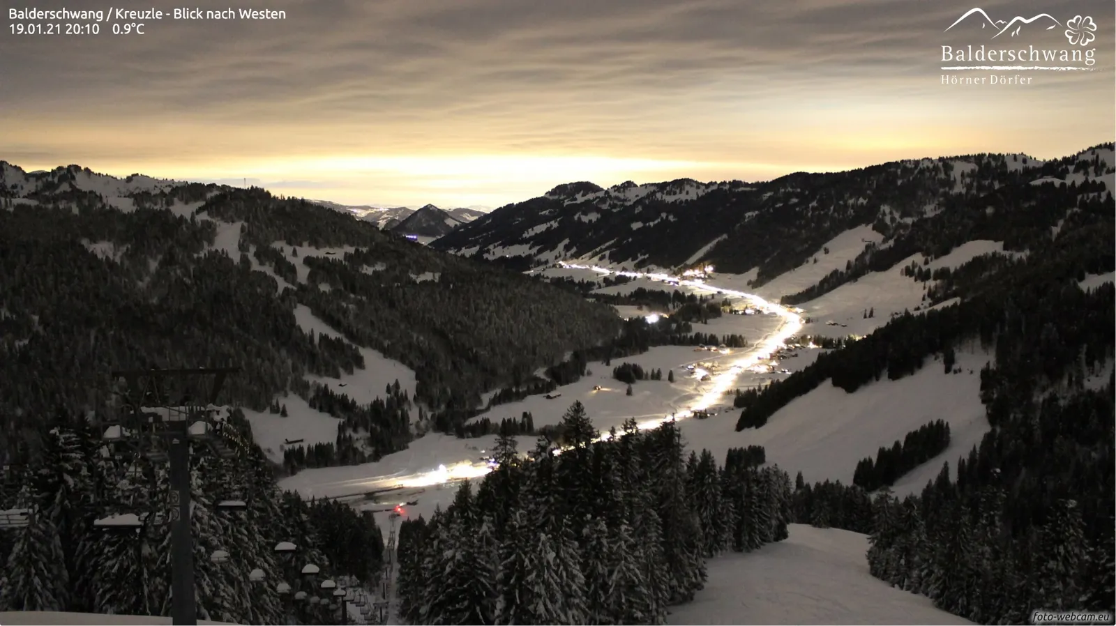Nachtaufnahme: verschneites Tal unter einer gelblich leuchtenden Wolkendecke, die Dorfstraße als helle Linie