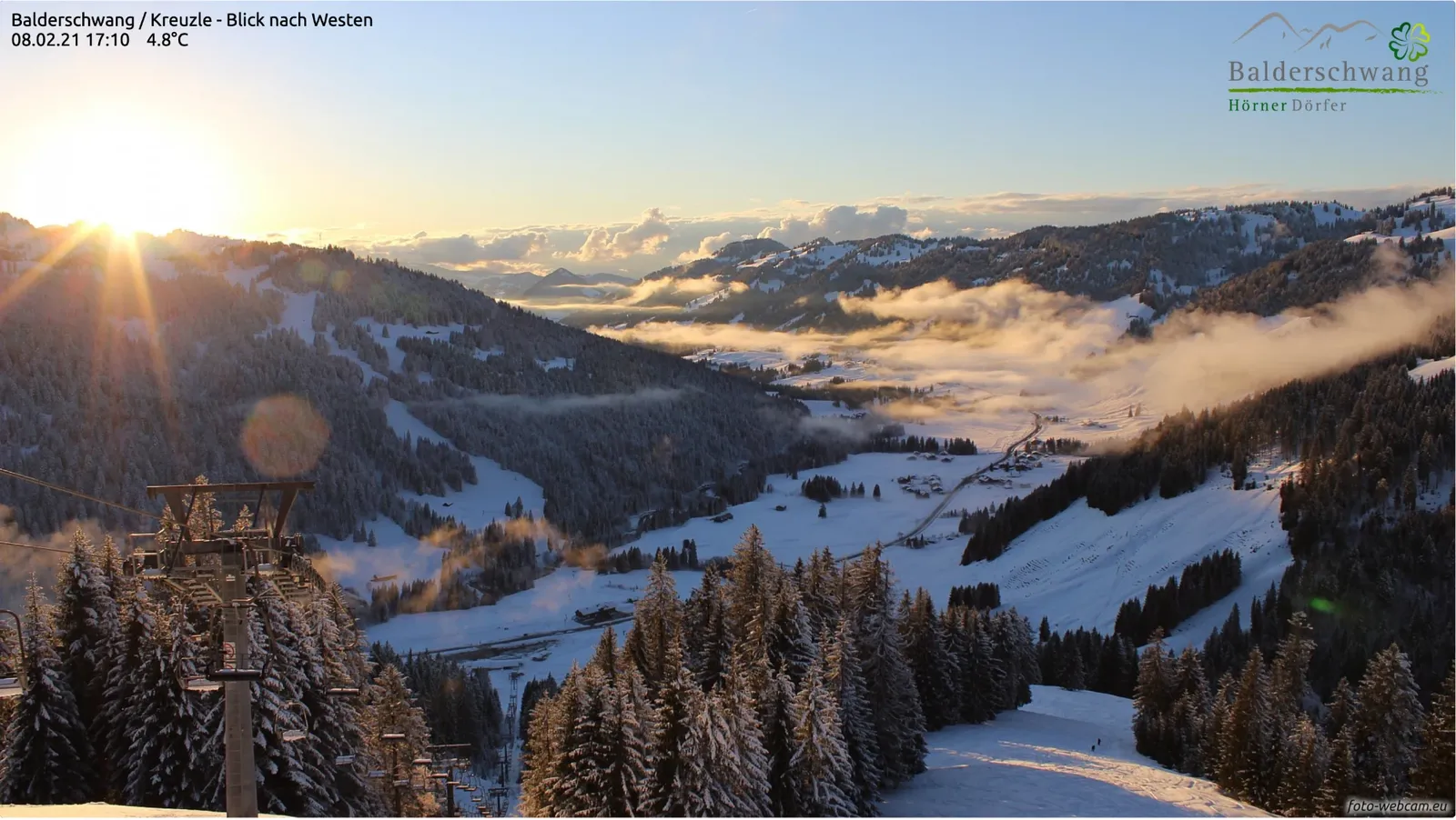 Winterlicher Sonnenuntergang über dem verschneiten Tal, Nebelschwaden liegen im Talgrund und leuchten golden