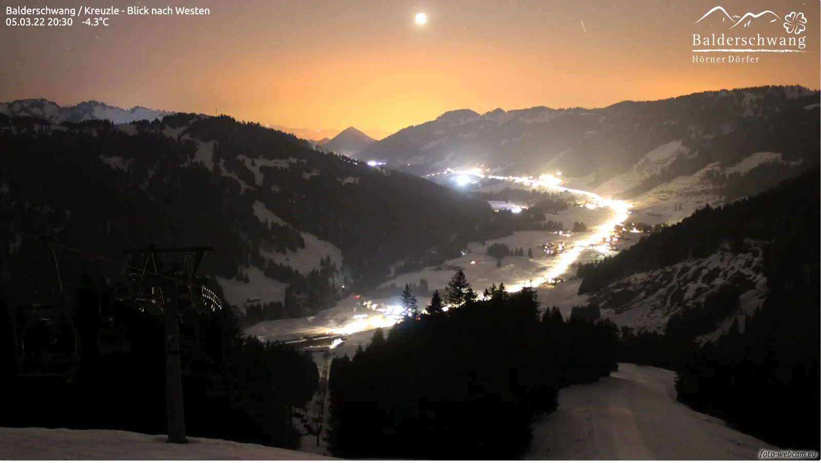 Der Mond über einem orange leuchtenden Nachthimmel, darunter das verschneite Tal mit der hellen Dorfstraße