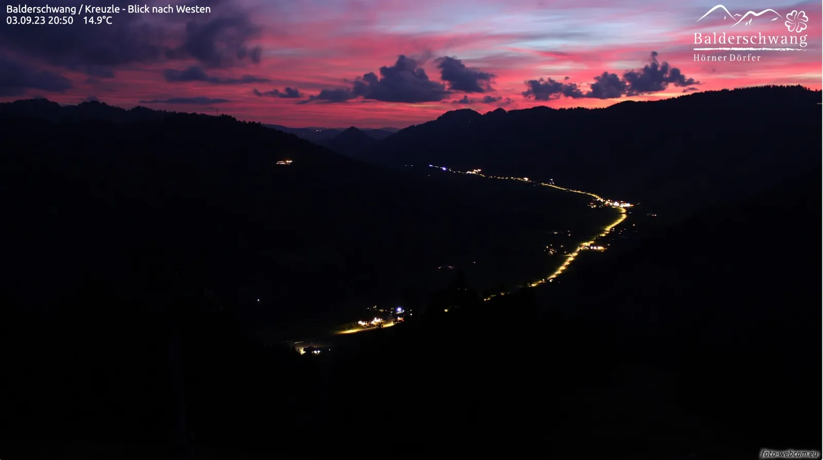 Leuchtend rosa und roter Abendhimmel mit dunklen Wolken, darunter das dunkle Tal mit der Lichterkette der Dorfstraße