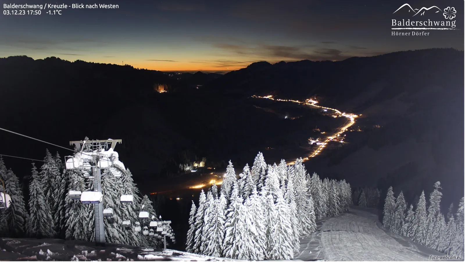Tief verschneite Tannen und Sessellift im Vordergrund, im dunklen Tal ein Lichterband, am Horizont ein orangefarbener Streifen