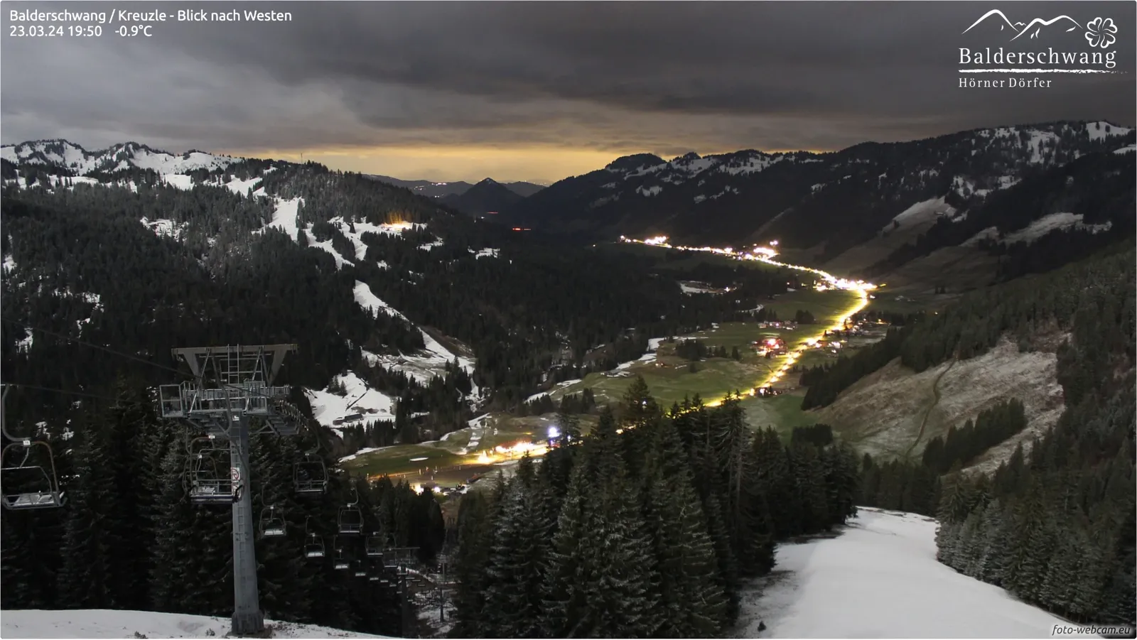 Dunkle Wolken über dem Tal, darunter ein gelber Lichtstreifen am Horizont und die hell beleuchtete Dorfstraße