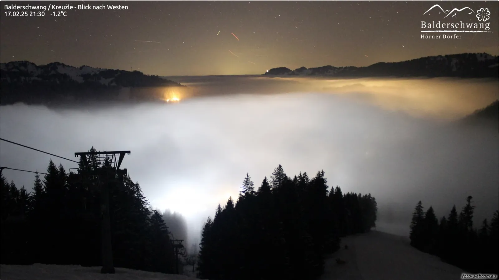 Sternenhimmel mit Lichtspuren über einem vom Dorf beleuchteten Nebelmeer