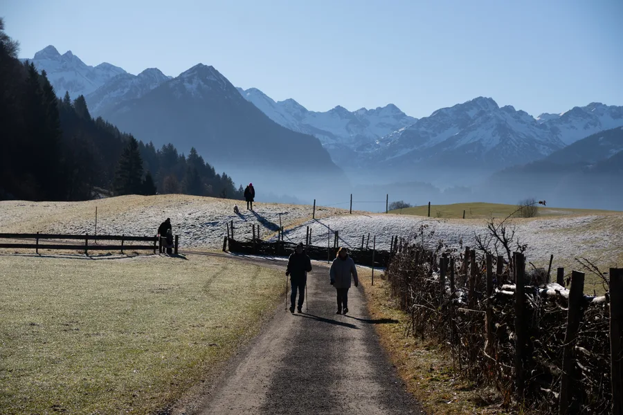 Spaziergänger auf einem Feldweg zwischen bereiften Wiesen, im Hintergrund verschneite Berge im Dunst